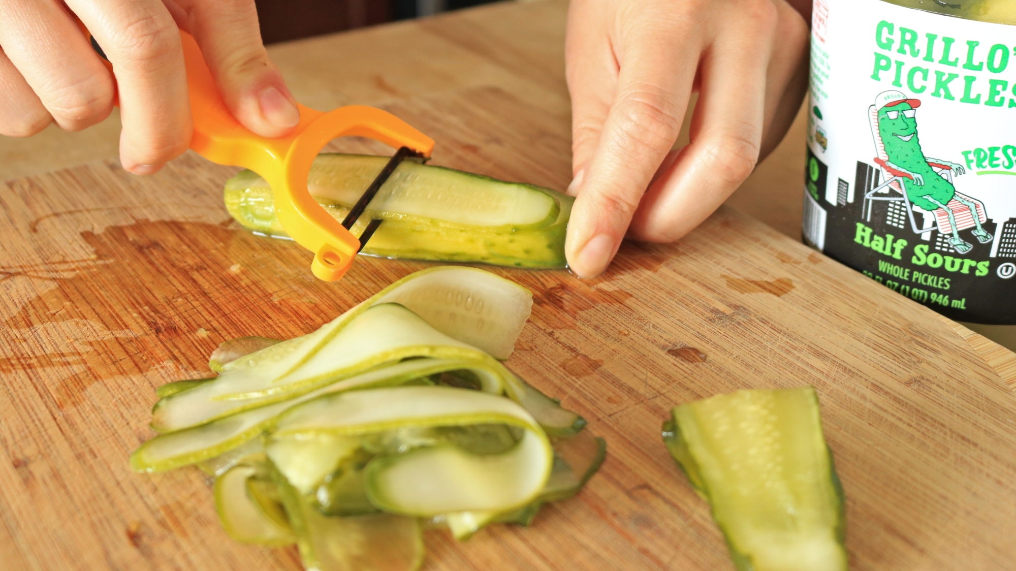 Hands using a vegetable peeler to shave a pickle.